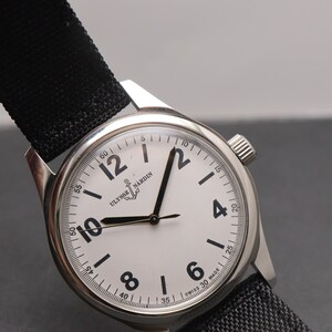May include: A stainless steel Ulysse Nardin wristwatch with a white dial, black Arabic numerals, and black hands. The watch features a black fabric strap, a silver-toned case, and the text Ulysse Nardin and Swiss Made printed on the watch face.