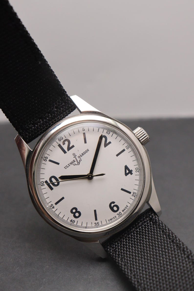 May include: A stainless steel Ulysse Nardin wristwatch with a white dial, black Arabic numerals, and black hands. The watch features a black fabric strap, a silver-toned case, and the text Ulysse Nardin and Swiss Made printed on the watch face.
