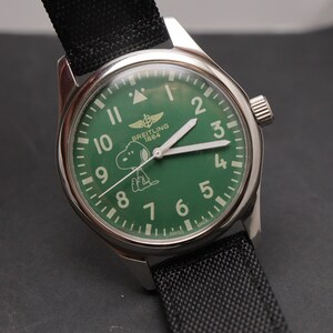 May include: A stainless steel wristwatch with a green dial featuring the Breitling 1884 logo and a white graphic of Snoopy. The watch has white hour markers, white hands, and a black textured fabric strap with a silver buckle and crown.