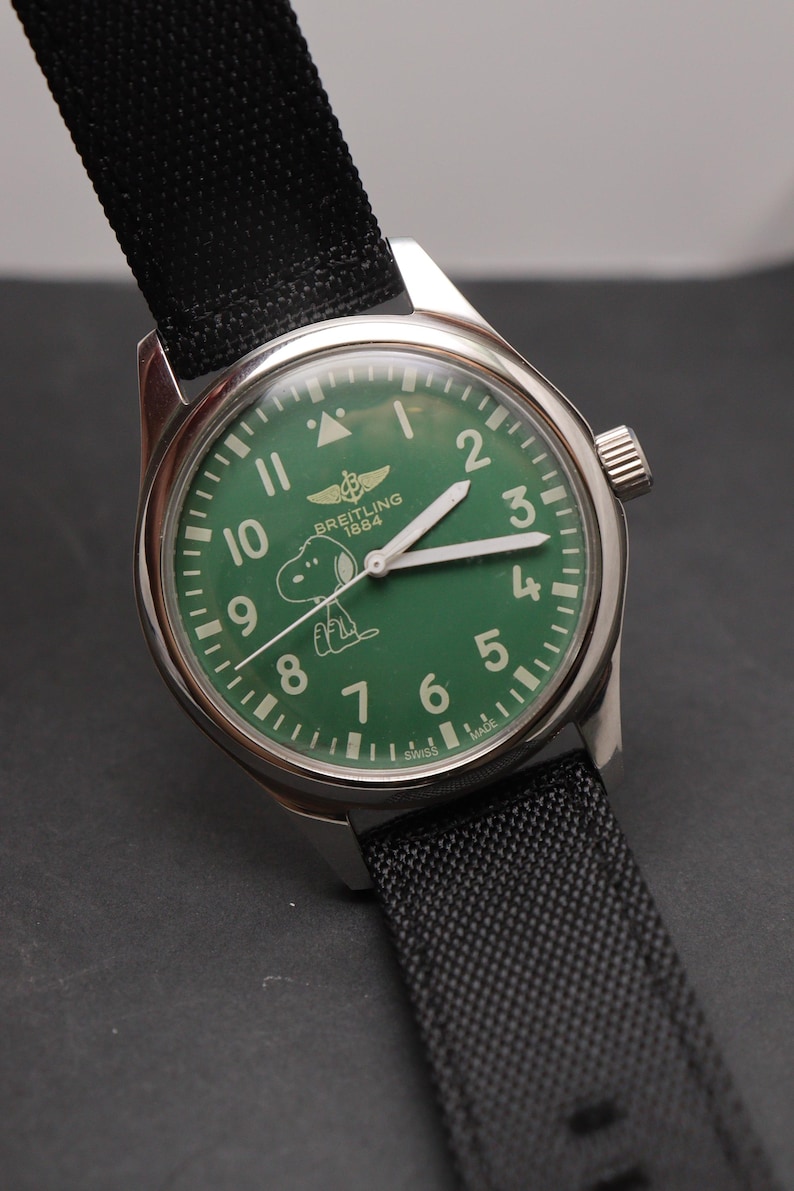 May include: A stainless steel wristwatch with a green dial featuring the Breitling 1884 logo and a white graphic of Snoopy. The watch has white hour markers, white hands, and a black textured fabric strap with a silver buckle and crown.