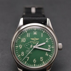 May include: A stainless steel wristwatch with a green dial featuring the Breitling 1884 logo and a white Snoopy graphic. The watch has white Arabic numerals, luminous hands, a triangle marker at the twelve o'clock position, and a black fabric strap.