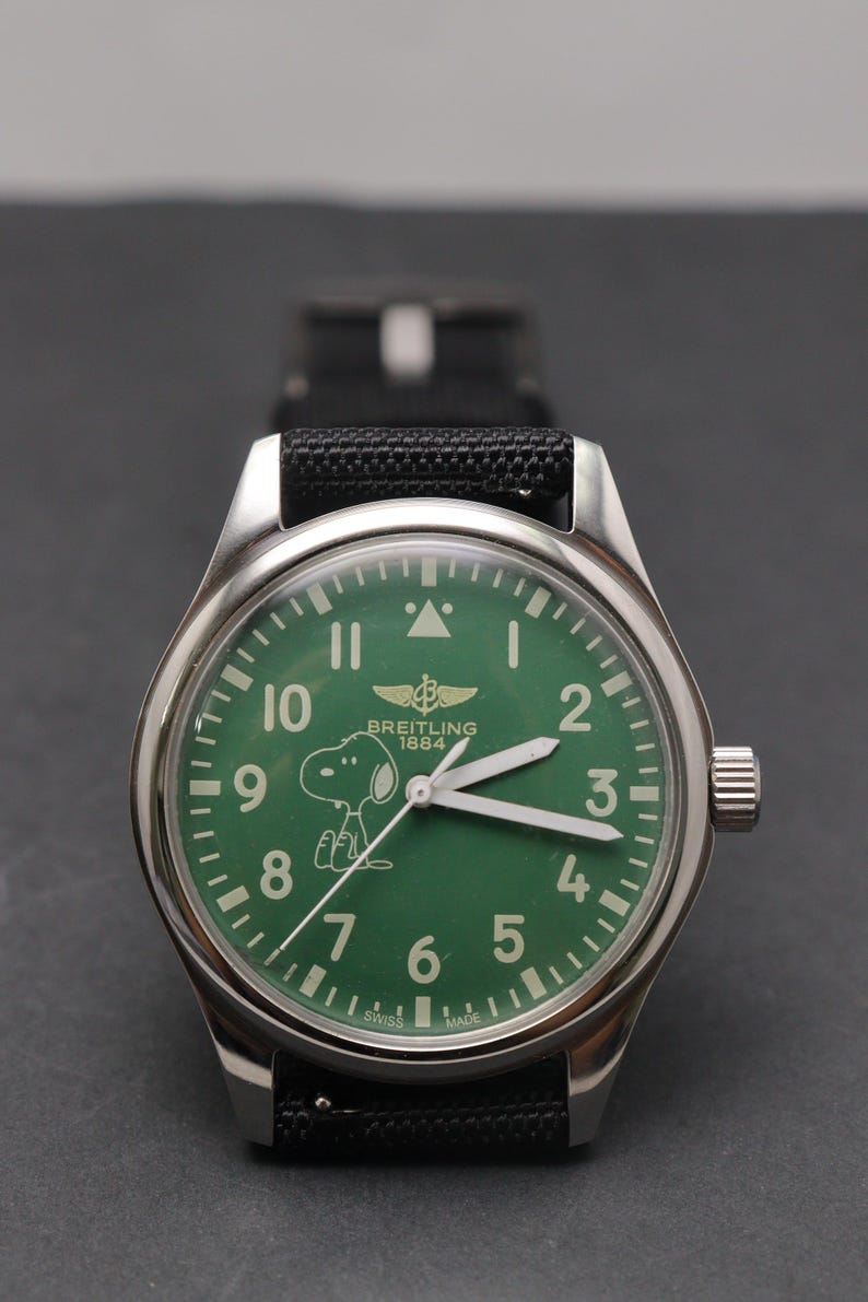 May include: A stainless steel wristwatch with a green dial featuring the Breitling 1884 logo and a white Snoopy graphic. The watch has white Arabic numerals, luminous hands, a triangle marker at the twelve o'clock position, and a black fabric strap.