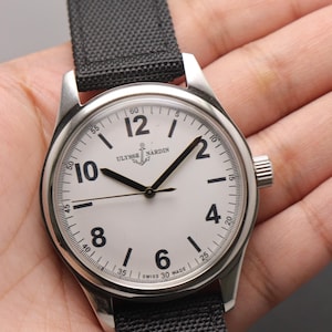 May include: A stainless steel wristwatch with a white dial, black Arabic numerals, and black hands. The dial features the Ulysse Nardin logo with an anchor icon and the text Swiss Made. It has a black fabric strap with a textured weave pattern.