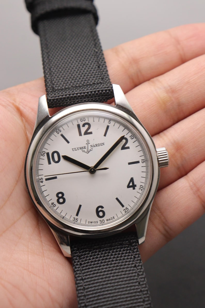 May include: A stainless steel wristwatch with a white dial, black Arabic numerals, and black hands. The dial features the Ulysse Nardin logo with an anchor icon and the text Swiss Made. It has a black fabric strap with a textured weave pattern.