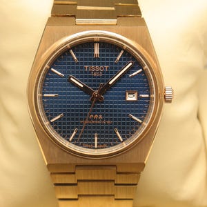 May include: A gold-tone stainless steel wristwatch with a blue waffle-patterned dial. The watch features gold-tone hour markers, hands, and a date window at the three o'clock position. The dial displays TISSOT 1853, PRX, POWERMATIC 80, and SWISS MADE.