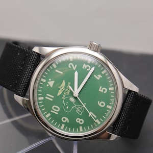 May include: A stainless steel wristwatch with a green dial featuring white Arabic numerals, a Breitling 1884 logo, and a graphic of Snoopy. The watch has white hands, a black fabric strap, and the text Swiss Made printed at the bottom of the dial.