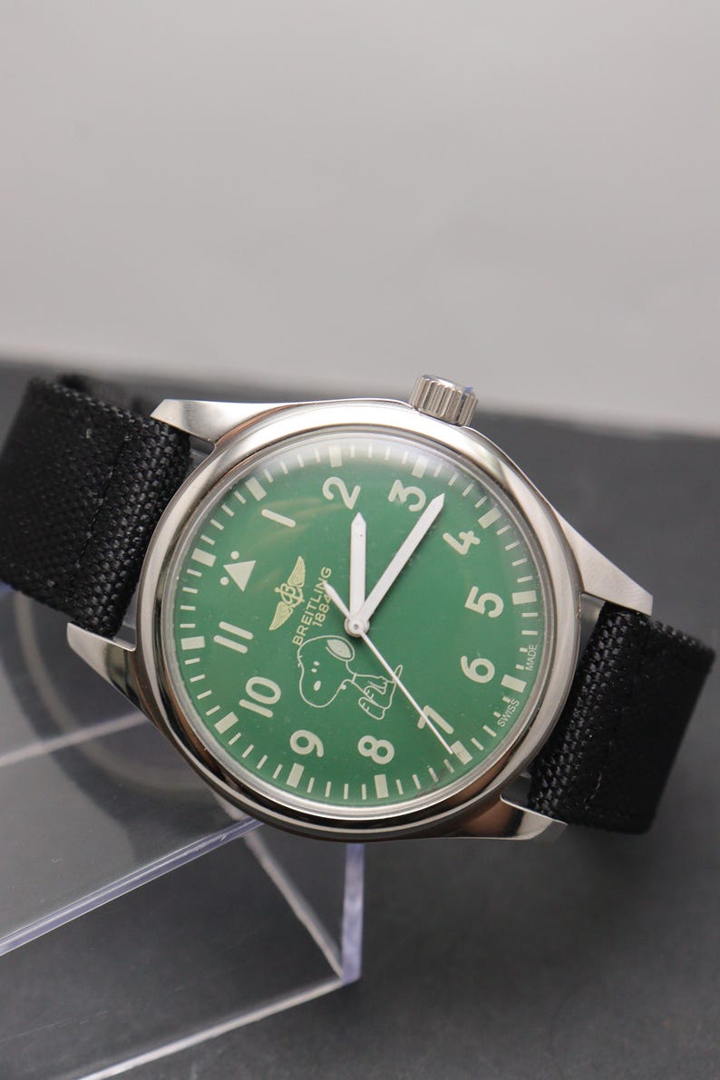 May include: A stainless steel wristwatch with a green dial featuring white Arabic numerals, a Breitling 1884 logo, and a graphic of Snoopy. The watch has white hands, a black fabric strap, and the text Swiss Made printed at the bottom of the dial.