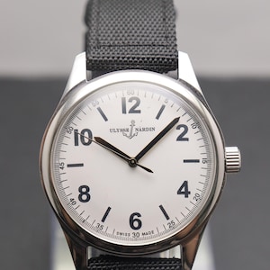 May include: A silver-tone wristwatch with a white dial, black Arabic numerals at the 2, 4, 6, 8, 10, and 12 positions, and black hands. The dial features the text ULYSSE NARDIN with an anchor logo and SWISS MADE. It has a black fabric strap.