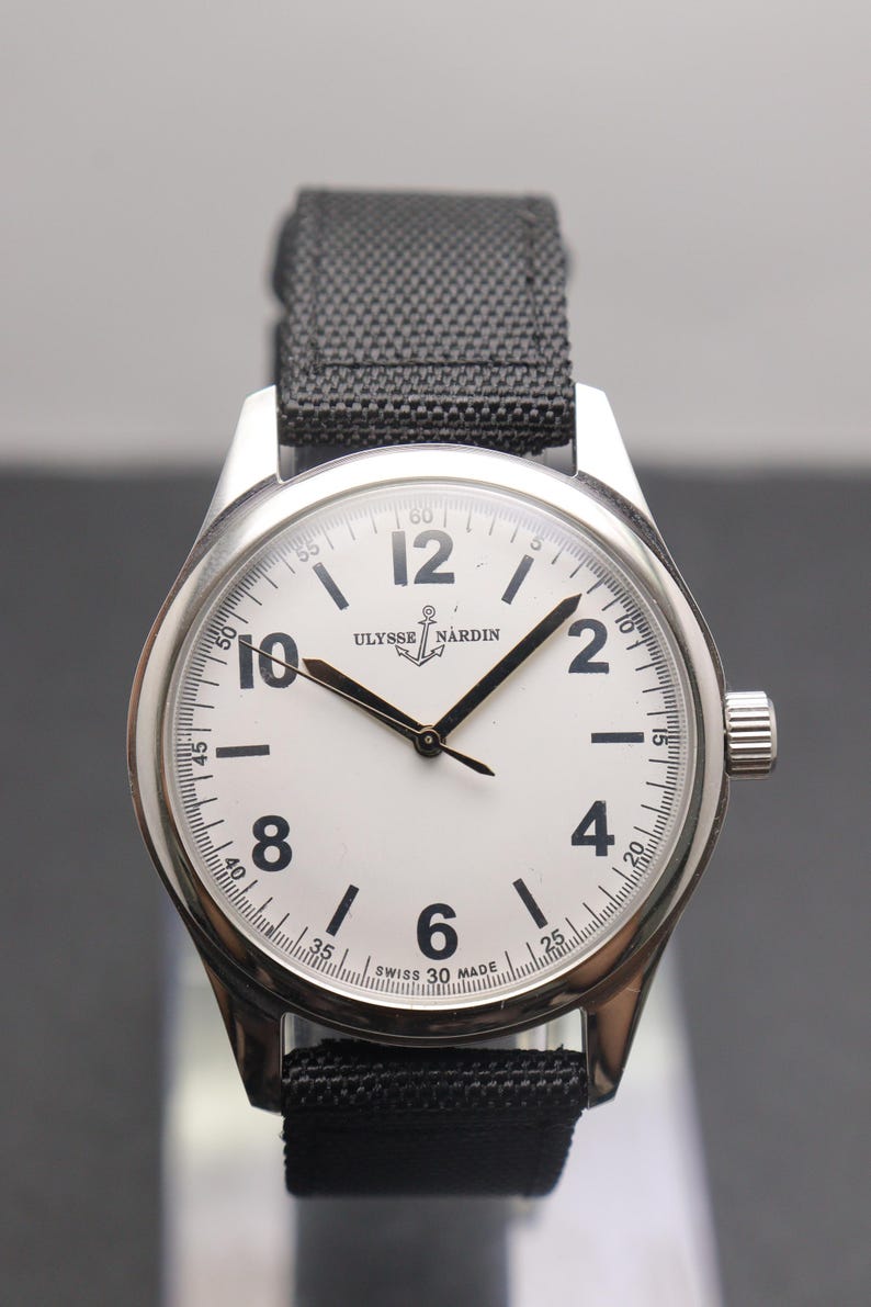 May include: A silver-tone wristwatch with a white dial, black Arabic numerals at the 2, 4, 6, 8, 10, and 12 positions, and black hands. The dial features the text ULYSSE NARDIN with an anchor logo and SWISS MADE. It has a black fabric strap.