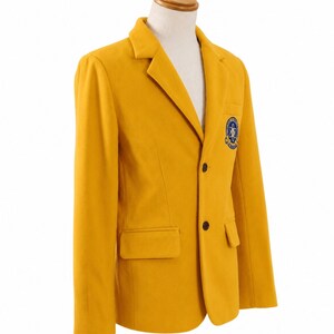 Puede incluir: Blazer amarillo mostaza con solapas de muesca, dos botones negros y un parche en el pecho. El blazer tiene dos bolsillos delanteros y se muestra en un maniqu&iacute;. El parche presenta un dise&ntilde;o azul y blanco.