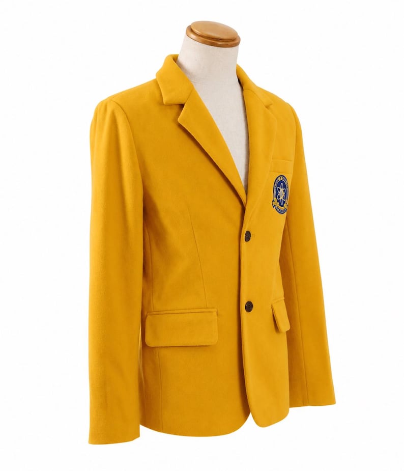 Puede incluir: Blazer amarillo mostaza con solapas de muesca, dos botones negros y un parche en el pecho. El blazer tiene dos bolsillos delanteros y se muestra en un maniqu&iacute;. El parche presenta un dise&ntilde;o azul y blanco.