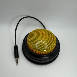 May include: A black and yellow button switch with a textured, yellow, circular top. The button is connected to a black cable with a 3.5mm audio jack. The base of the button is black and round.