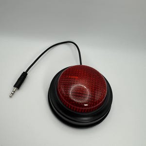 Può includere: Un pulsante rotondo rosso con una superficie strutturata e una base nera. Un cavo nero si estende dal pulsante, terminando con un jack audio da 3,5 mm. Probabilmente un dispositivo di assistenza tecnologica.