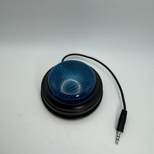 May include: A blue, textured button with a black base and a black cable ending in a 3.5mm jack. The button is round and dome-shaped, with a grid-like pattern on its surface. The background is plain white.