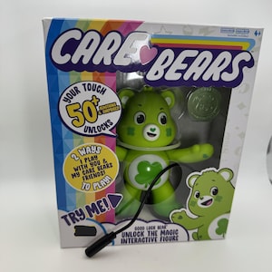May include: A Good Luck Bear interactive figure from the Care Bears series, packaged in a colorful box. The green bear has a four-leaf clover on its belly and a smiling face. The box features text including "50+ Reactions & Surprises" and "Unlock the Magic".