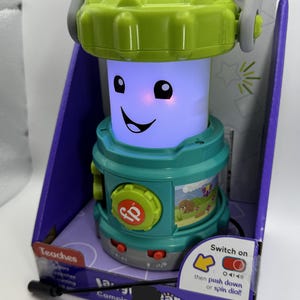 Może przedstawiać: Zielono-turkusowa latarnia zabawka z uśmiechniętą twarzą z przodu. Latarnia ma uchwyt i pokrętło z napisem "Switch on" i "Camping Fun Lantern". Opakowanie wskazuje, że zabawka uczy piosenek i zwrotów i jest przeznaczona dla dzieci w wieku 6-36 miesięcy.