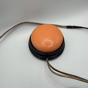 May include: A round, orange push-button switch with a smooth, domed surface set into a black circular base. The device features a thin, transparent-coated wire extending from the side, terminating in a standard black 3.5mm audio jack connector.