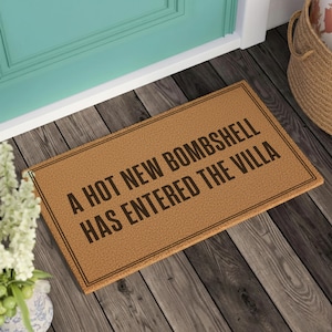 May include: A rectangular tan coir doormat with a black border and the text A HOT NEW BOMBSHELL HAS ENTERED THE VILLA printed in black capital letters. The mat features a coarse, natural fiber texture and is positioned on a dark wooden floor.