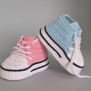 May include: A pair of handmade crochet baby sneakers. One sneaker is pink and white, the other is blue and white. Both have black stripes on the soles and white laces. The sneakers are designed to look like classic high-top shoes.