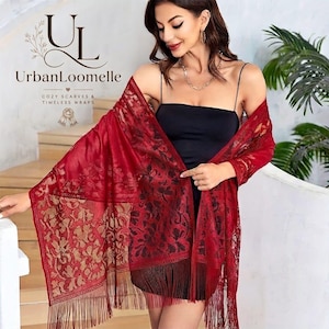 May include: A deep red rectangular shawl featuring an intricate floral lace pattern with metallic gold accents and long fringe trim along the edges. The fabric is semi-sheer, showcasing a detailed botanical design throughout the entire wrap.