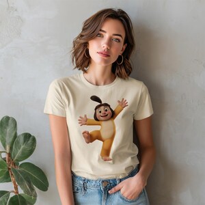 May include: A cream-colored short-sleeve t-shirt featuring a centered graphic of a cartoon character with dark hair in a ponytail, wearing a yellow onesie, with arms outstretched and one leg lifted in a joyful, dancing pose.