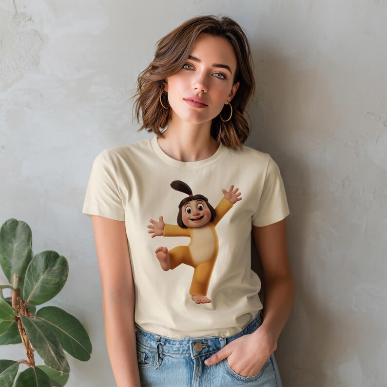 May include: A cream-colored short-sleeve t-shirt featuring a centered graphic of a cartoon character with dark hair in a ponytail, wearing a yellow onesie, with arms outstretched and one leg lifted in a joyful, dancing pose.