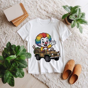 May include: A white short-sleeved t-shirt featuring a colorful graphic of a clown with rainbow-colored hair driving a tan off-road vehicle. The clown has a red nose and is waving one hand. The vehicle has black tires and a brown interior.