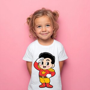 May include: A white short-sleeved t-shirt featuring a graphic print of a cartoon character with black hair, wearing a red and yellow superhero costume with a star emblem on the chest, saluting with a white-gloved hand against a solid pink background.