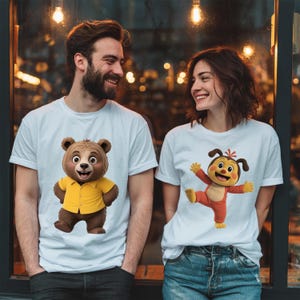 May include: Two white t-shirts featuring cartoon characters. The left shirt displays a brown bear wearing a yellow button-down shirt. The right shirt features a brown and tan cartoon creature with floppy ears wearing a red outfit with arms outstretched.