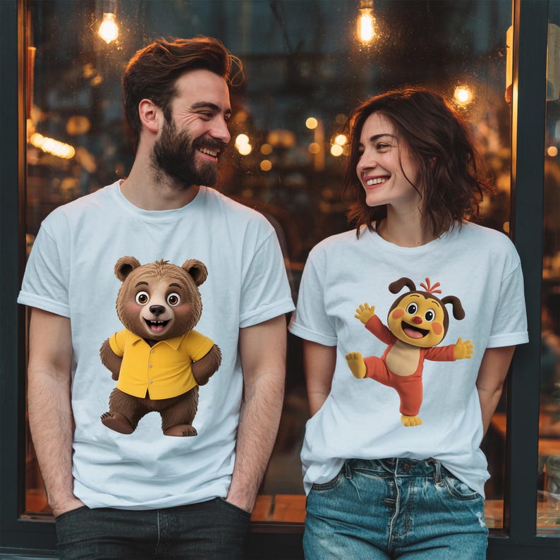 May include: Two white t-shirts featuring cartoon characters. The left shirt displays a brown bear wearing a yellow button-down shirt. The right shirt features a brown and tan cartoon creature with floppy ears wearing a red outfit with arms outstretched.