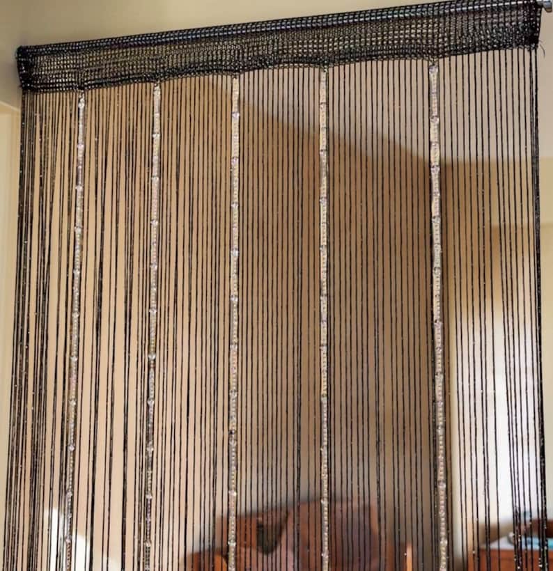 May include: A black string curtain featuring a solid woven header at the top. The curtain is composed of numerous thin, vertical black strands interspersed with several vertical columns of clear, reflective, rectangular beads that catch the light.