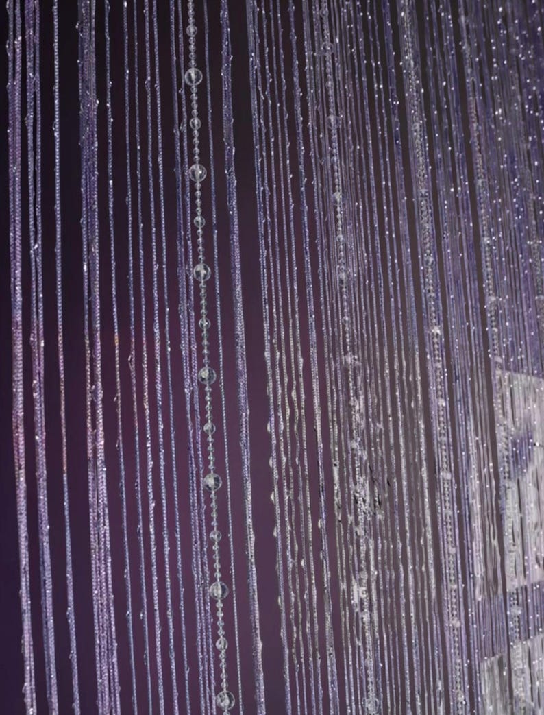 May include: A string curtain featuring numerous vertical strands of thin, light-reflecting material. The strands are interspersed with small, clear, spherical beads that catch the light, creating a shimmering effect against a dark background.