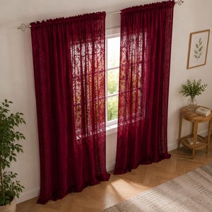 May include: A pair of deep red, semi-sheer curtains featuring an intricate, repeating floral lace pattern. The curtains are hung on a silver-toned metal rod with decorative finials, covering a window and allowing soft light to filter through.