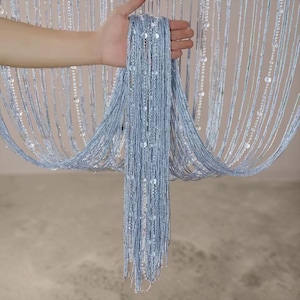 May include: A light blue string curtain featuring vertical strands of shimmering metallic thread interspersed with clear, round beads. The curtain is composed of numerous thin, hanging lines that create a dense, textured, and reflective appearance.