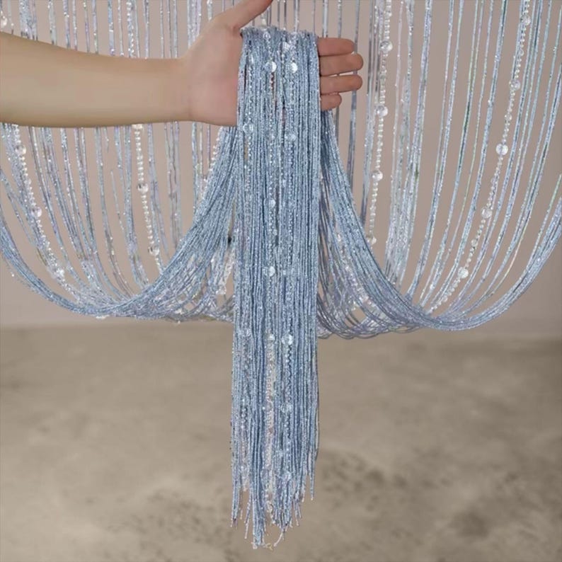 May include: A light blue string curtain featuring vertical strands of shimmering metallic thread interspersed with clear, round beads. The curtain is composed of numerous thin, hanging lines that create a dense, textured, and reflective appearance.