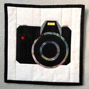 Könnte beinhalten: Eine gesteppte Kamera in Schwarzweiß mit einem bunten Objektiv. Die Kamera ist von einem schwarzen Rahmen und einem weißen Hintergrund umgeben.