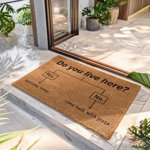 May include: A rectangular brown coir doormat featuring a black printed flowchart graphic. The text reads: Do you live here? with two branches: Yes, leading to welcome home, and No, leading to come back with pizza. The mat has a coarse texture.