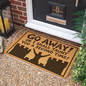 May include: A rectangular tan coir doormat featuring a dark brown silhouette of a castle and a person with raised arms. The text printed in dark brown reads: GO AWAY! OR I SHALL TAUNT YOU A SECOND TIME! The mat has a dark brown border.