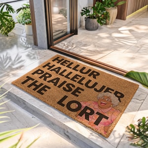 May include: A rectangular tan coir doormat featuring the black text HELLUR HALLELUJER PRAISE THE LORT. The mat includes a colorful graphic of an older person with gray hair wearing glasses and a pink floral shirt, positioned in the bottom right corner.
