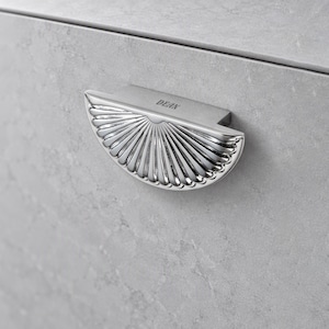 May include: A semi-circular silver cabinet pull with a fan-like ribbed pattern and the word DEAN engraved on the top flat edge. The hardware is mounted on a light gray stone surface with a subtle mottled texture and a horizontal seam.