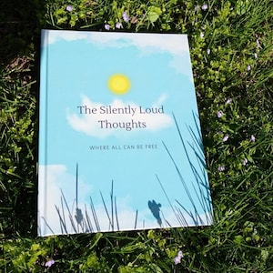 May include: A hardcover book titled "The Silently Loud Thoughts" with a blue sky and yellow sun design on the cover. The book is lying on a bed of green grass with small purple flowers.