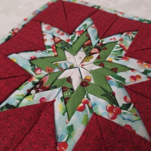 May include: A handmade, quilted star-shaped trivet. The trivet features a central star design in green fabric with floral and berry patterns, surrounded by red triangles. The edges are also red, creating a festive, holiday-themed look.