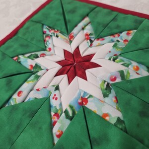 May include: A handmade, square quilted trivet with a starburst design. The center star is red, surrounded by white and fabric with holly and berry patterns. Green triangles form the outer points, with a red border.