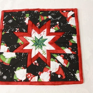 May include: A square quilted potholder featuring a central star pattern in white and green, surrounded by a black fabric background printed with gnomes, red hats, and white snowflakes. The entire piece is bordered with a solid red fabric trim.