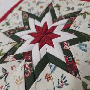 May include: A handmade Christmas star-shaped quilted trivet. The trivet features a central red star, surrounded by white, green, and cream-colored fabric panels. The cream panels are decorated with holly and other festive motifs, and the edges are trimmed with red.