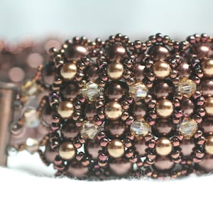 May include: A close-up of a brown and gold beaded bracelet with a woven pattern. The bracelet is made with small, round beads and has a delicate, intricate design.