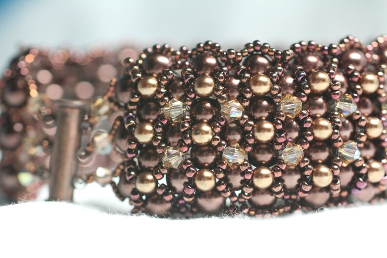 May include: A close-up of a brown and gold beaded bracelet with a woven pattern. The bracelet is made with small, round beads and has a delicate, intricate design.