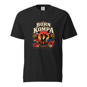 May include: Black t-shirt with a colorful graphic. The graphic features the words "Born to Kompa" above a silhouette of a dancing couple, palm trees, drums, and flowers. The design is set against a sunset background.