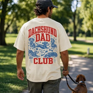 Peut inclure: Un t-shirt couleur cr&egrave;me avec un motif rouge et bleu au dos. Le texte indique DACHSHUND DAD CLUB et Life is better with doxies. Le design comprend des illustrations de teckels dans diverses positions, une balle, un os et une gamelle.