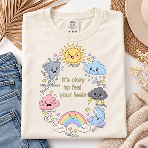 Puede incluir: Una camiseta de color crema con un gráfico circular de varios personajes relacionados con el clima con caras. El texto en el centro dice "it's okay to feel your feels", rodeado por un sol, nubes, un tornado, un arcoíris y una luna con etiquetas.